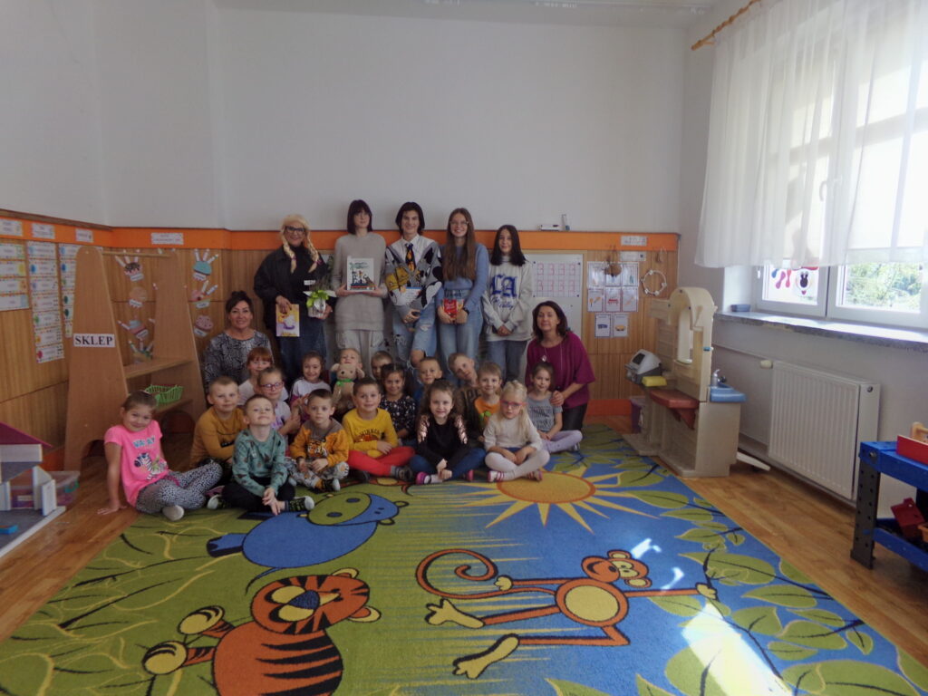 Zdjęcie grupowe. Na dywanie siedzą dzieci z paniami i młodzieżą zaproszona do głośnego czytania.