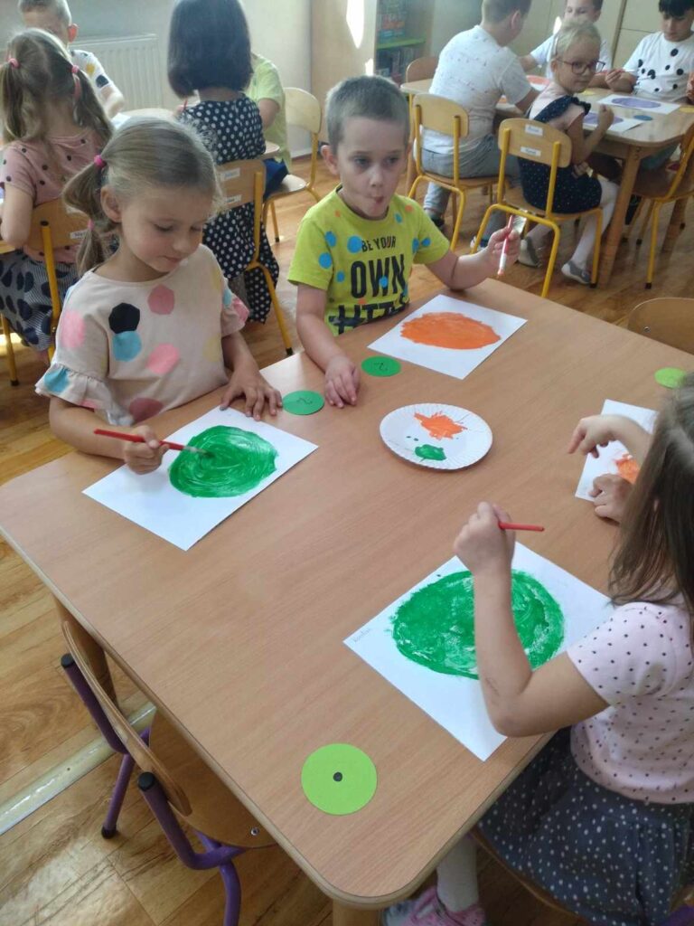 Dzieci przy stoliku podczas pracy plastycznej. dzieci malują farbami dużą kropkę. 
