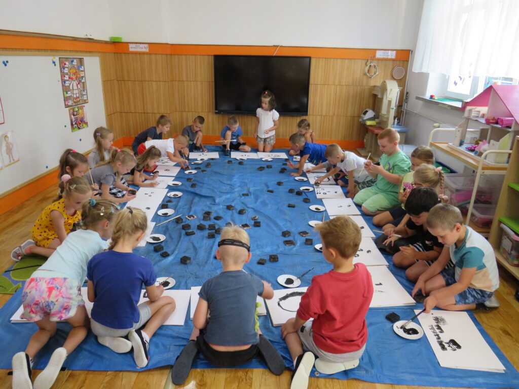 Zdjęcie grupowe. Dzieci siedzą wokół rozłożonej na dywanie plandeki. Malują czarną farbą swój autoportret. 