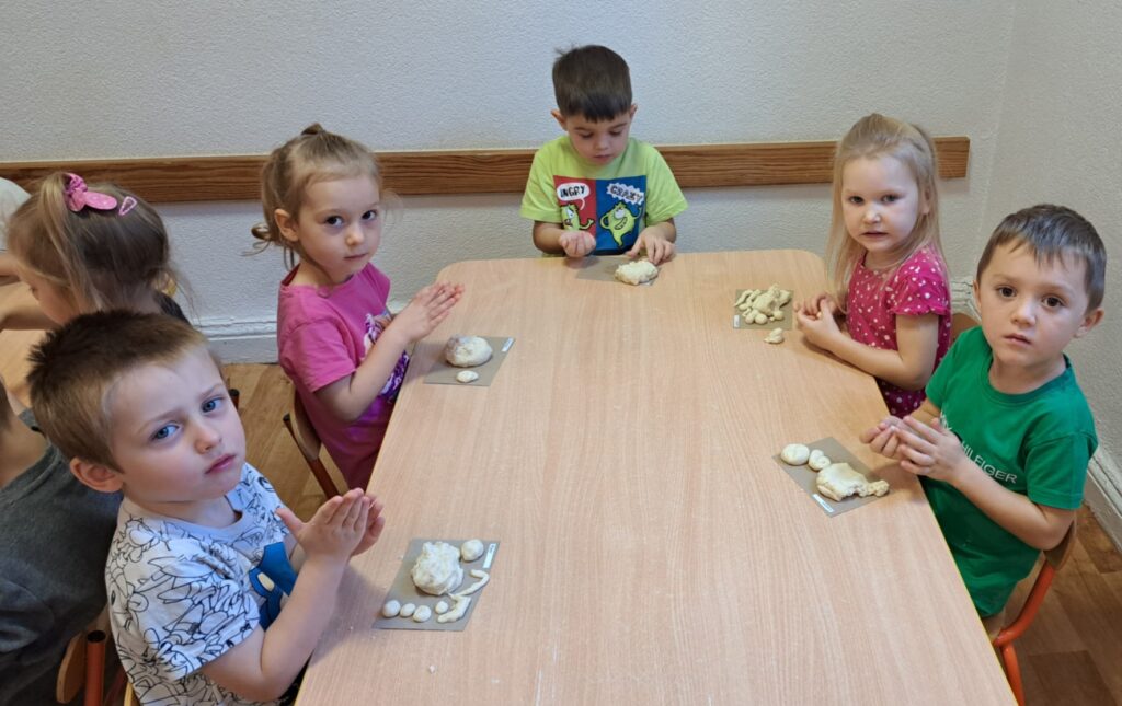 W sali przedszkolnej przy stolikach siedzą dzieci. W rączkach kleją chlebki, bułeczki z masy solnej.