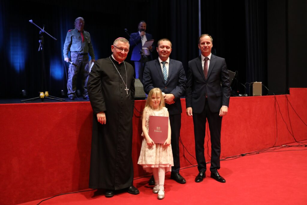 Zdjęcie zrobione podczas odbioru nagrody w konkursie plastycznej. Zdjęcie przedstawia dziewczynkę z biskupem, burmistrzem i starostą miasta Sławno. 