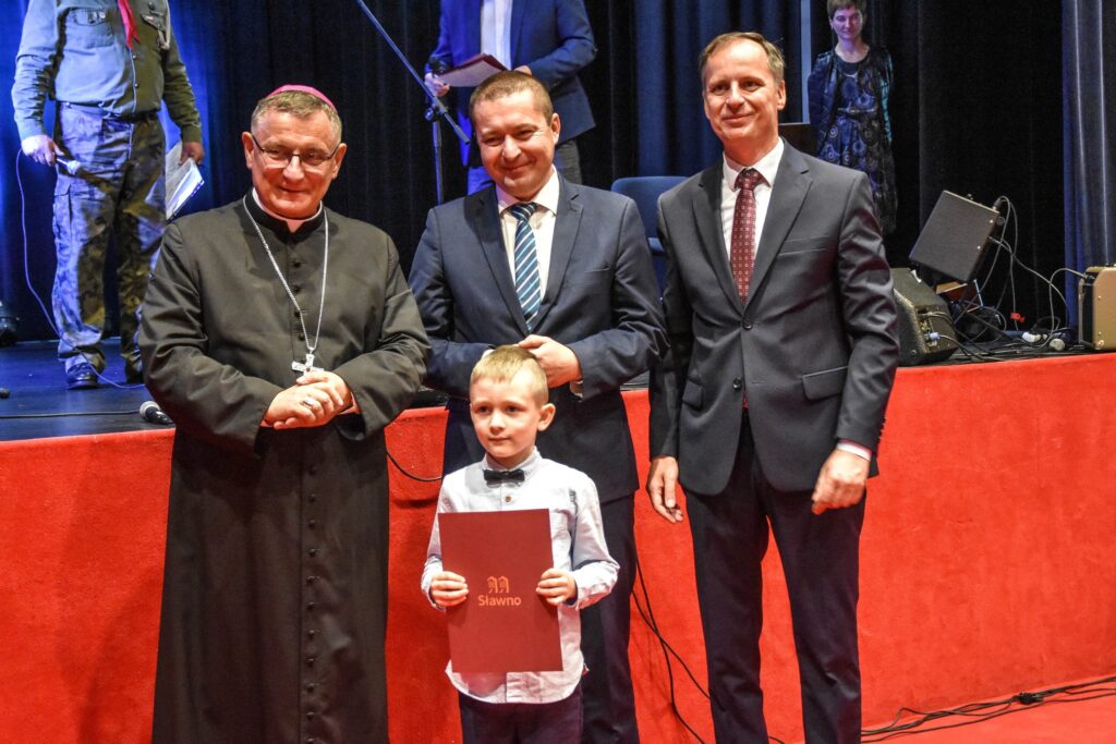 Zdjęcie zrobione podczas odbioru nagrody w konkursie plastycznej. Zdjęcie przedstawia chłopca z biskupem, burmistrzem i starostą miasta Sławno. 
