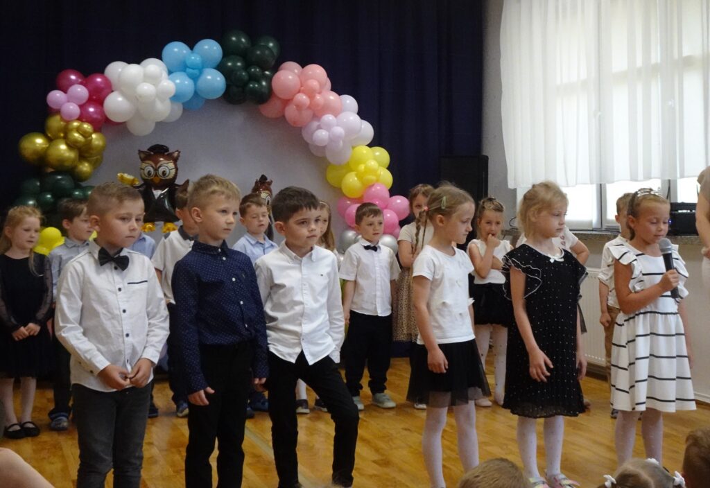 Dzieci ustawione w szeregu. Jedna dziewczynka trzyma w rączce mikrofon. 