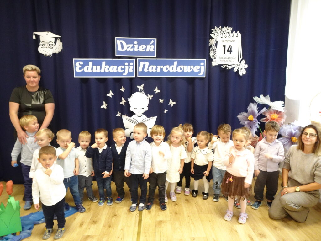 Zdjęcie grupowe. Dzieci w galowych strojach na tle dekoracji z napisem Dzień Edukacji Narodowej. 