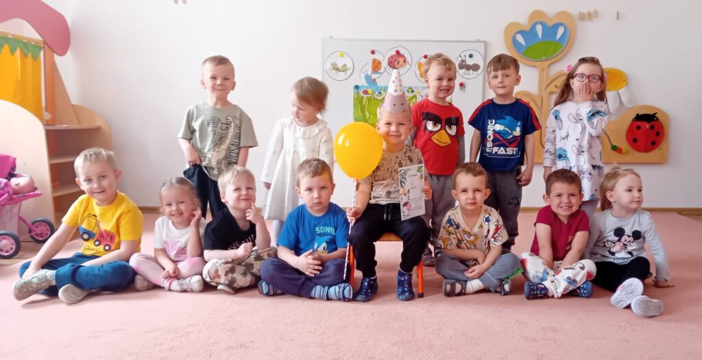 Zdjęcie grupowe dzieci z okazji urodzin. W środku, na krzesełku siedzi chłopiec w urodzinowej czapeczce.