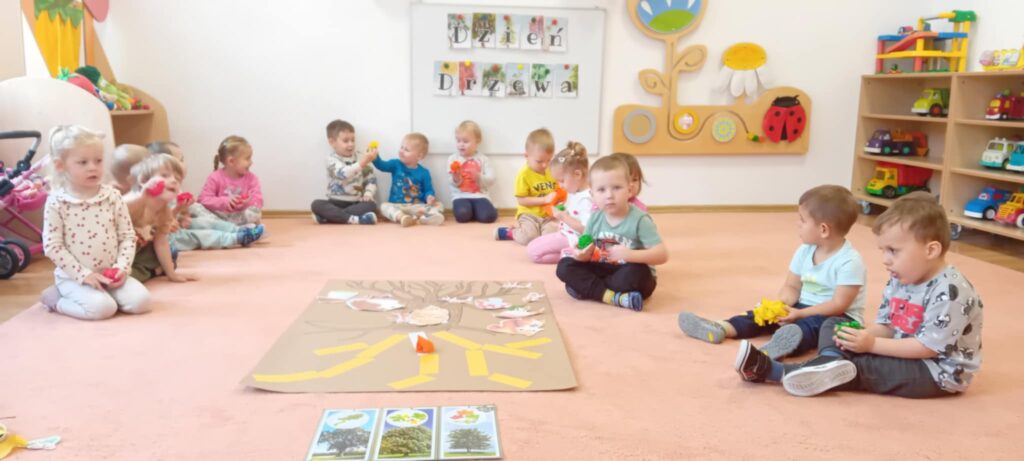 Dzieci siedzą na dywanie. W rączka trzymają kolorową bibułę, którą zgniatają w kulki. 