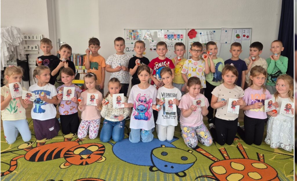 NA DYWANIE SIEDZĄ I STOJĄ DZIECI W RĘKACH TRZYMAJĄ PREZENCIK I KINDER JAJKO