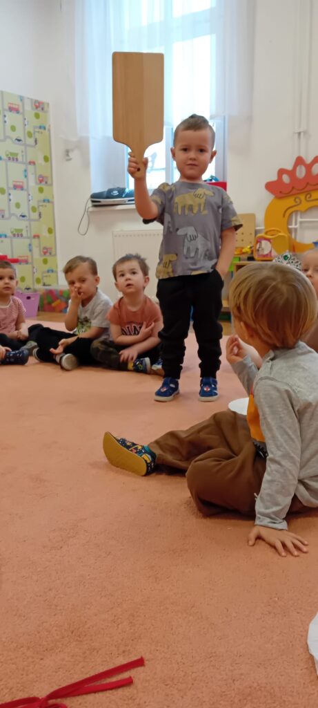 Dzieci podczas zajęć na dywanie. Jeden z chłopców prezentuje deskę do krojenia.