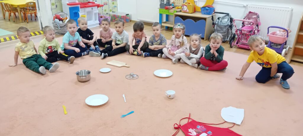 Dzieci podczas zajęć na dywanie. Na środku dywanu rozłożone są kuchenne przedmioty.
