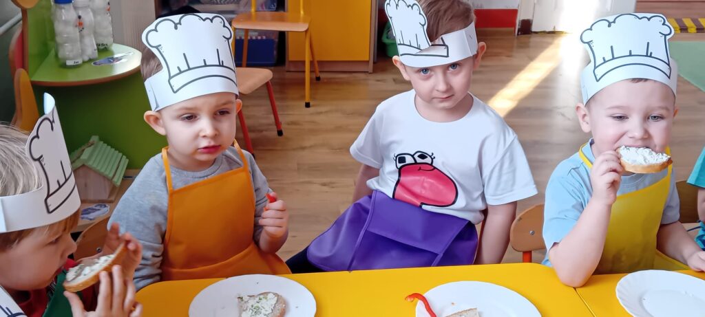 Dzieci przy stoliku podczas jedzenia własnoręcznie zrobionej kanapki z twarożkiem.