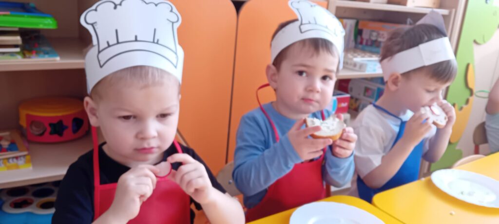 Dzieci przy stoliku podczas jedzenia własnoręcznie zrobionej kanapki z twarożkiem.