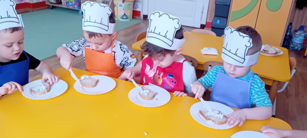 Dzieci przy stoliku podczas smarowania pieczywa twarożkiem.